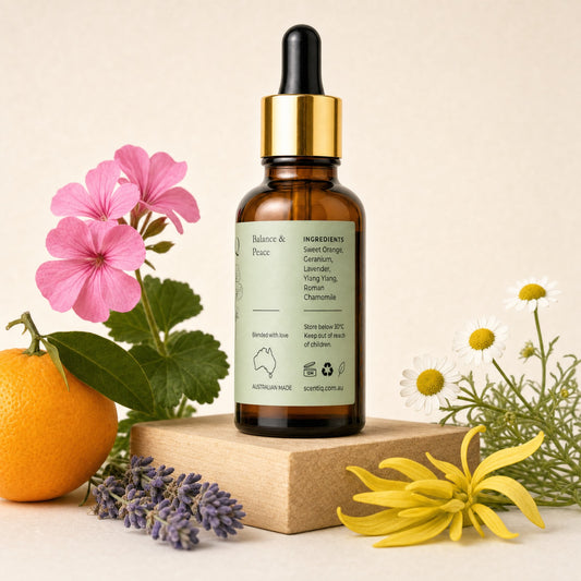 An amber glass dropper bottle labeled 'Balance & Peace' stands on a wooden block, surrounded by botanical props including pink flowers, a lemon, lavender, white daisies, and a yellow flower against a warm beige background.