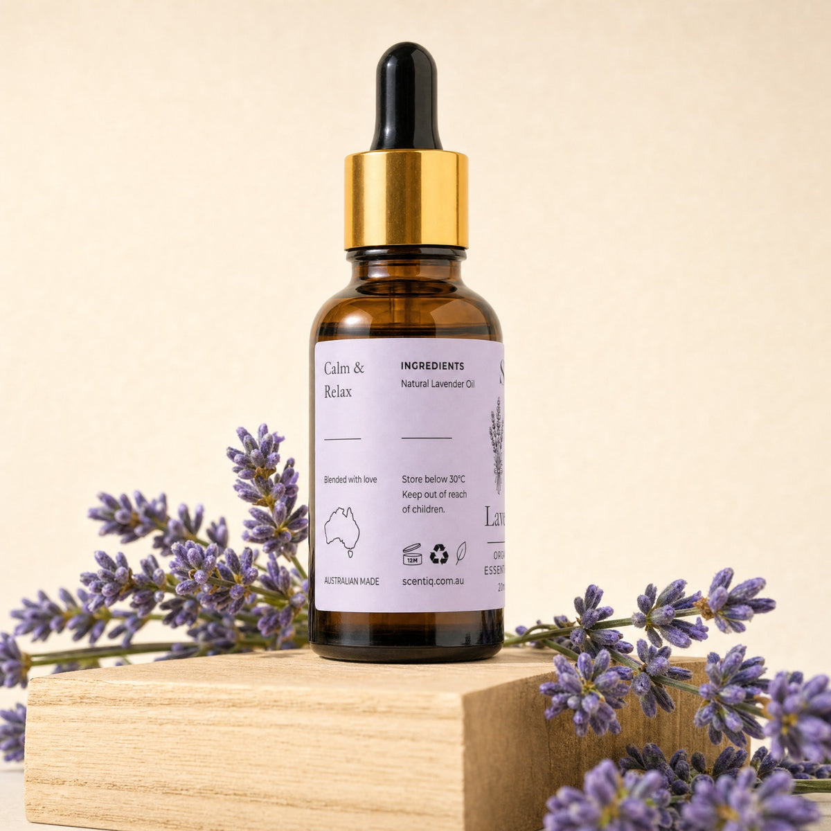 An amber glass dropper bottle of lavender oil with a gold collar and black dropper cap, standing on a wooden block with lavender sprigs around it against a warm beige background.