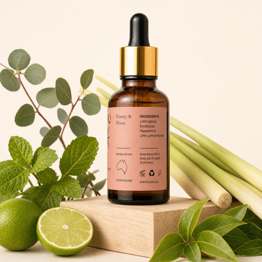 An amber glass dropper bottle with a gold collar and a peach-pink label reading 'Energy & Focus' and listing lemongrass, eucalyptus, peppermint, and lime. The bottle is styled on a wooden block with fresh green limes, mint leaves, and eucalyptus stems against a warm cream background.